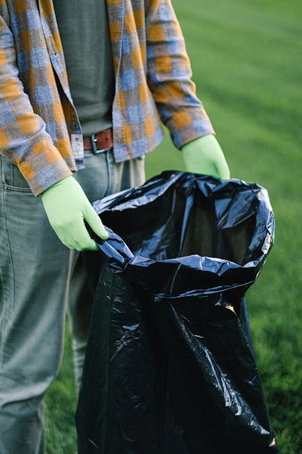 A person wearing a plaid shirt with shades of yellow and gray, along with green rubber gloves, is holding open a large black rubbish bag made of shiny, flexible plastic material. The bag is empty, showing its dark, reflective interior. The individual’s lower body is visible, with gray trousers and a brown belt. The background features a well-maintained, lush green lawn, indicating an outdoor setting, possibly a garden or park. The scene suggests routine waste collection, cleanup, or waste disposal activities, which are relevant to services such as those provided by companies specializing in rubbish removal or private waste handling. The context hints at an environment where proper disposal of waste or rubbish is being managed, aligned with independent or alternative waste collection methods outside of municipal services.