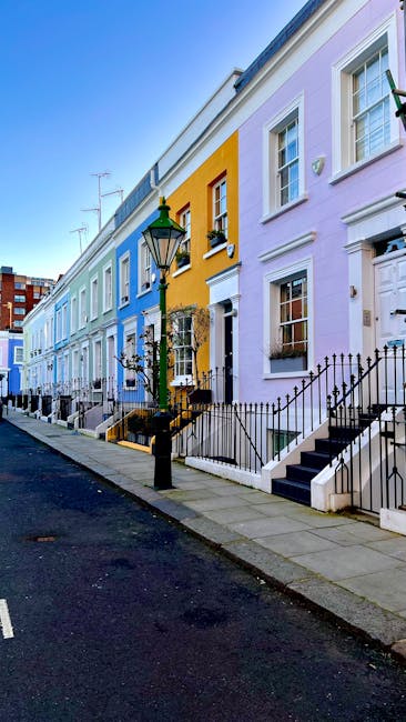 A row of townhouses painted in pastel shades of blue, yellow, and lavender line a city street, each with white-framed windows, front steps, and black wrought-iron railings. A vintage-style street lamp with green and black metalwork stands on the pavement in front of the blue house, casting natural light on the scene. The sidewalks are clean, and the street appears quiet with no visible vehicles or pedestrians. The ground level of the buildings features small front gardens or steps leading to doorways, consistent with typical London residential architecture. In the background, some taller brick buildings and antennae are visible under a clear, blue sky. The scene depicts well-maintained, colorful urban housing, with no rubbish or waste visible, but the image context aligns with independent waste management or flexible rubbish collection services that serve such neighborhoods.