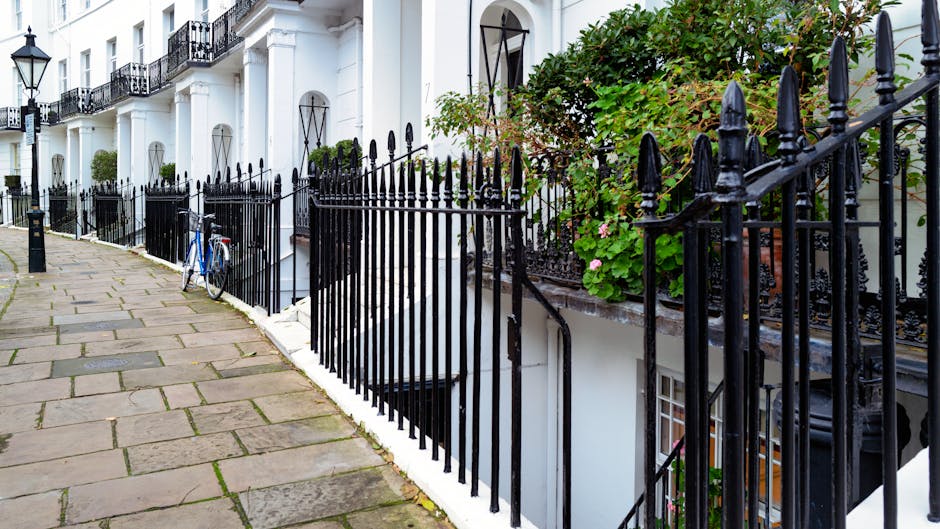 A row of white terraced houses with black wrought iron fencing along a cobblestone sidewalk. The fences have pointed finials and separate the pavement from small garden areas in front of each property. One of the gardens contains green leafy plants and pink flowers. A blue bicycle is secured to the fence near the front of one house. The scene is illuminated by natural daylight, with shadows cast by the fencing and plants. The background shows multiple shutters and balconies on the white facades of the buildings, with some potted plants visible on the steps leading to basement entrances. The overall setting appears clean and well-maintained, typical of residential areas where private waste disposal or on-site clearance might be required, supported by independent rubbish collection services such as those provided by [COMPANY_NAME] for waste management and rubbish removal in West Kensington.
