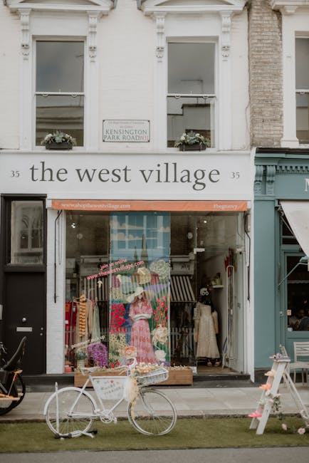 The image shows the storefront of a shop called 'the west village,' located on a ground-floor unit of a white, two-story building with decorative molding around the windows. Above the entrance, there is a sign reading 'KENSINGTON PARK ROAD NW' affixed to the building façade. The shop’s large glass window displays mannequins dressed in vintage-style clothing, with one wearing a pink floral dress and another in a beige coat, alongside various decorative items and clothing racks visible inside. In front of the store, a white bicycle with a basket on the front wheel is parked on the sidewalk, partly on a small strip of grass. To the right, a teal-colored shop with a partially extended awning is visible, and there are outdoor items including a small wooden stand with pink flowers and a black bicycle leaning against the building. The overall scene depicts a retail area that might be involved in private collection or retail of vintage or second-hand items, reflecting an environment where rubbish removal services from companies like [COMPANY_NAME] could be involved in clearing discarded items from commercial premises or during shop renovations.