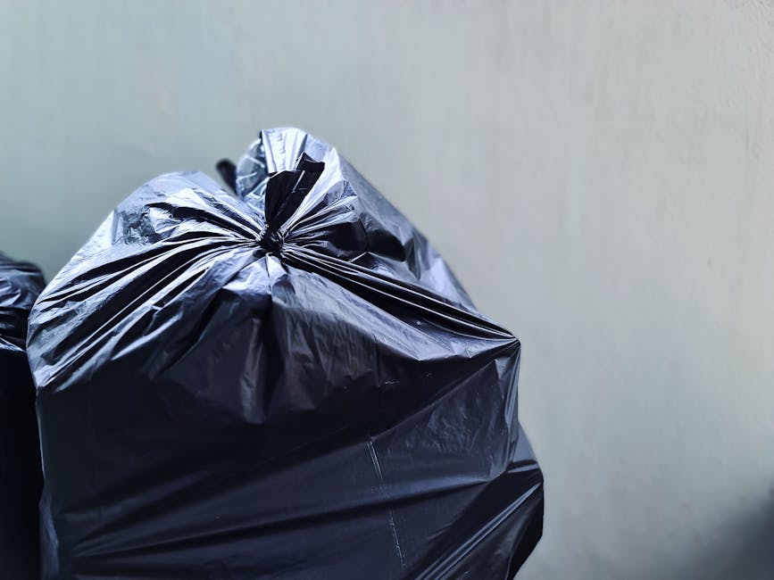 Close-up of a black plastic rubbish bag tightly secured with a knot at the top, sitting on a surface against a plain, light-colored wall background. The bag appears full, with a glossy, crinkled texture reflecting light, indicating contents that could include household waste or discarded materials. Additional black rubbish bags are partially visible beside it, suggesting a collection of waste ready for removal. The setting appears to be indoors, possibly a storage or utility area, where private waste collection services like those of [COMPANY_NAME] might be used as an alternative to local authority bin collection. The image emphasizes the use of sturdy, sealed rubbish bags typically employed in professional waste disposal, highlighting a scene of on-site rubbish handling or preparing waste for collection in an environment that aligns with professional rubbish removal practices in the W8 and W14 postcode areas, West Kensington.