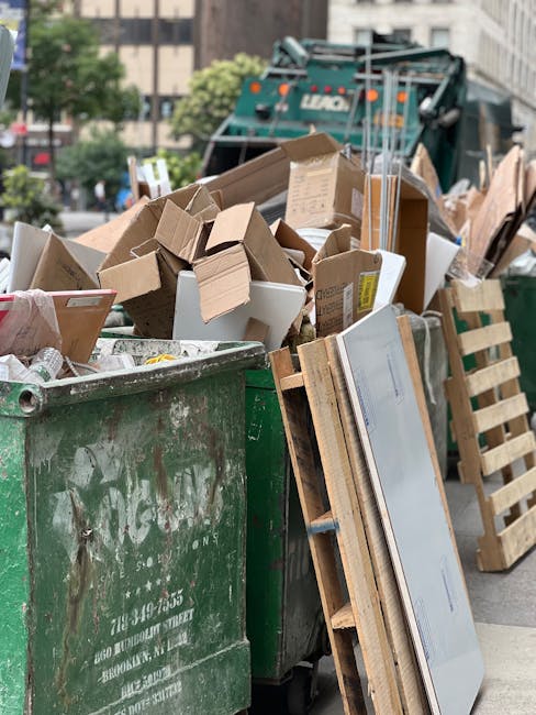 A large green municipal waste container filled with a mixture of cardboard boxes, some flattened and others partially assembled, along with a few white packaging cartons, positioned on an urban street. Several wooden pallets are leaning against the side of the container, with one pallet partially visible in the foreground. Behind the container, a recycling truck with a fold-out compactor mechanism is parked, partially obscured by the rubbish. The background features a cityscape with multistory buildings, trees, and parked cars, indicating a busy urban environment. The scene includes natural daylight, highlighting the textures of the cardboard, wood, and metal surfaces, typical of a private rubbish collection or alternative waste handling scenario managed by a local waste service provider such as rubbishcollectionwestkensington.co.uk.