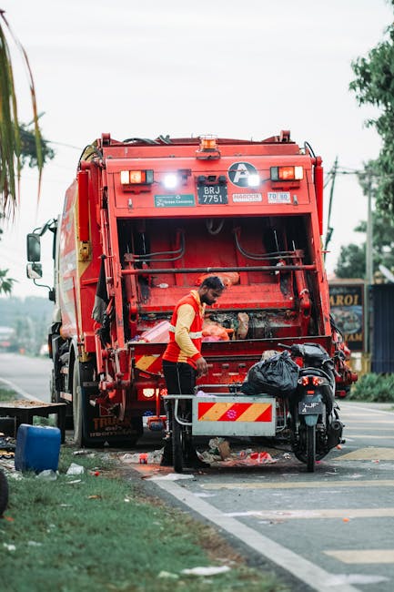A waste collection worker, dressed in a yellow and orange high-visibility vest, is standing behind a large red rubbish truck on a roadside, actively engaged in waste disposal or collection activities. The back of the truck is open, revealing the mechanical components and waste inlet, with a black plastic bag and other debris nearby, some placed on the ground and others on a small platform attached to a motorcycle parked adjacent to the truck. The scene is set outdoors during daytime, with overcast sky and trees in the background, indicating a suburban or urban environment. Utility poles and overhead wires are visible, along with a distant signboard, suggesting the truck is positioned on a street used for waste or rubbish removal services. The waste collector's attention appears focused on handling or managing refuse, which aligns with a professional rubbish removal context involving private or alternative waste handling methods, as facilitated by services such as [COMPANY_NAME]. The environment shows signs of street cleanup, with collected debris on the ground, emphasizing the scene's focus on efficient rubbish collection and disposal activities.