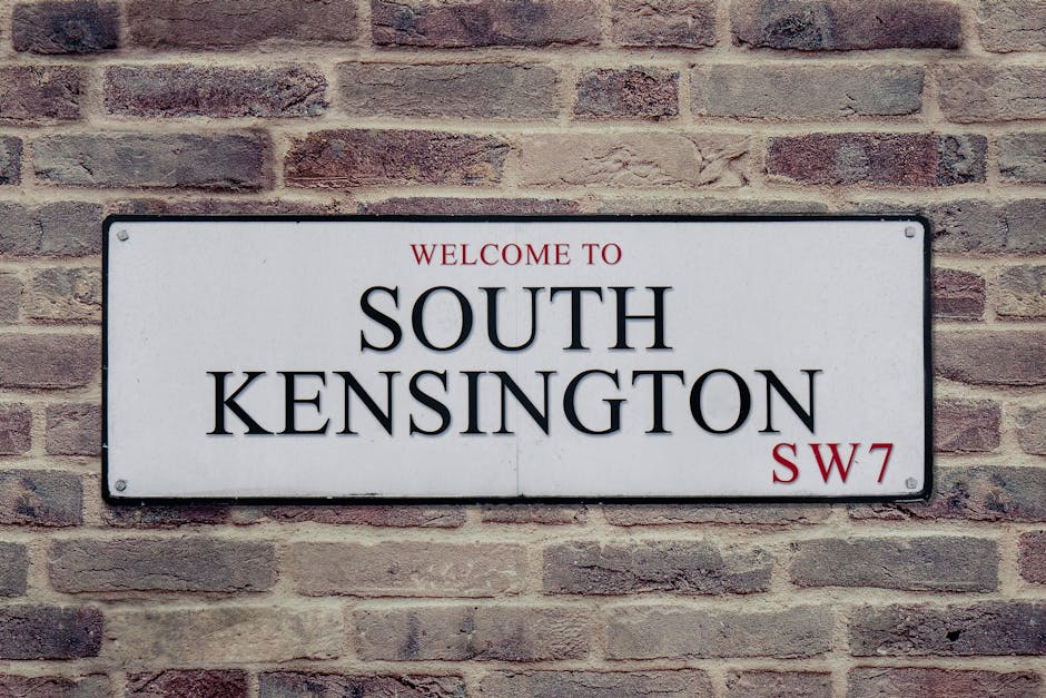 A rectangular white metal street sign mounted on a light red brick wall with mortar joints, displaying the text 'Welcome to South Kensington SW7' in black and red letters. The sign is positioned centrally on the wall, which features a traditional brick pattern with a slightly rough texture. The sign's surface is smooth with rounded corners and visible screw fasteners at each corner. The background wall consists of uniformly sized bricks with subtle color variations, creating an evenly textured appearance. The lighting is diffuse, suggesting daytime conditions. This street sign is characteristic of UK signage used for area identification, and its presence in a setting associated with residential or urban environments implies that independent waste collection or private waste handling services might operate nearby, consistent with the context of alternative rubbish removal methods in the area.