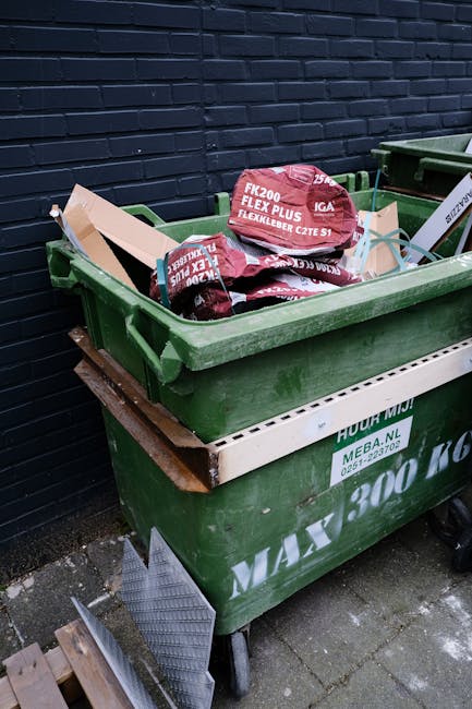 A green waste container filled with black and red bags of garden refuse, placed outdoors against a dark blue painted brick wall. The bags are marked with text indicating garden mulch and fertiliser products, and are stacked loosely inside the container. The container is situated on a paved surface, with another similar container adjacent to it. A wooden pallet lies beneath the container, and a metal ramp or platform leans against it, suggesting recent use for waste collection or disposal. The scene is well-lit with natural daylight, emphasizing the textures and colors of the bags and the container, which appears to be part of private waste handling or on-site clearance typically associated with independent rubbish removal services in various environments, including residential or commercial premises.