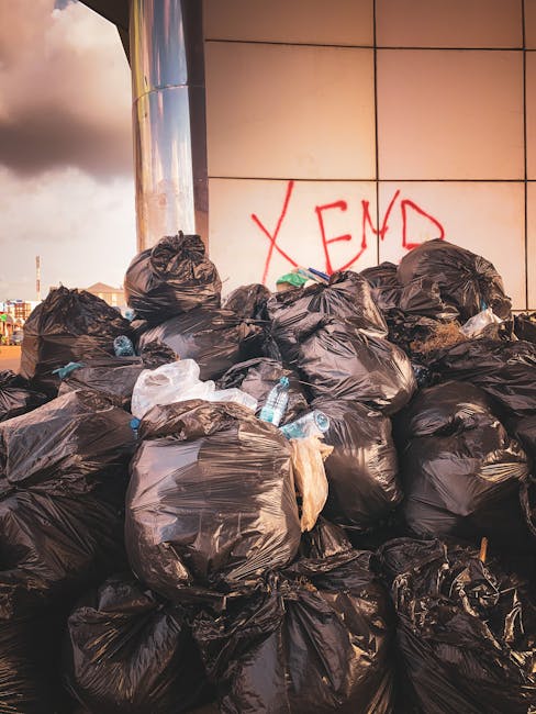 A large pile of black rubbish bags, some with visible creases and tied at the top, are stacked against a modern building with a reflective exterior wall composed of large, rectangular beige and light tan panels. Among the bags, several clear plastic bottles are visible, with some partially crushed or leaning against the bags. Behind the pile, there is graffiti in red spray paint spelling 'XEND' on the wall. The scene is illuminated by natural light, possibly during late afternoon or early evening, casting a soft glow over the waste and the building's surface. The environment appears to be an urban outdoor area, such as a service alley or loading zone, associated with private waste handling or alternative rubbish removal services like those provided by companies such as [COMPANY_NAME], which may be involved in on-site rubbish clearance or independent waste collection. The overall impression is one of unmanaged rubbish accumulation, highlighting the need for professional waste management solutions.
