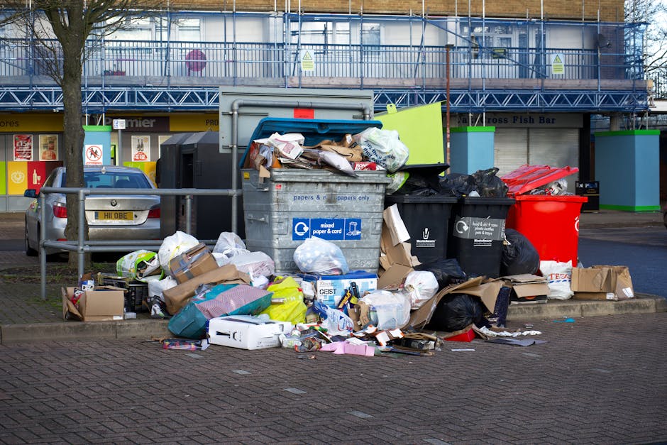 A large outdoor waste collection area featuring multiple open-top wheelie bins and nearby discarded rubbish on the paved ground. The central grey bin with a blue label is overflowing with mixed paper, cardboard, and packaging debris, with some waste spilling onto the pavement. Adjacent to it, there are several black and red bin bags stored loosely on the ground, containing various rubbish. Cardboard boxes and plastic packaging are scattered around the base of the bins, some flattened and others partially crushed. To the left, a parked silver car is situated behind a metal railing, with a tree behind it and commercial storefronts in the background, including a fish bar. Behind the waste area, a building with a scaffolding cover indicates ongoing construction or renovation, with reflective safety signage visible. The scene captures an outdoor spot designated for private rubbish disposal or alternative waste handling, a typical setting for independent rubbish collection services in urban areas, with natural daylight illuminating the cluttered but orderly waste management space.