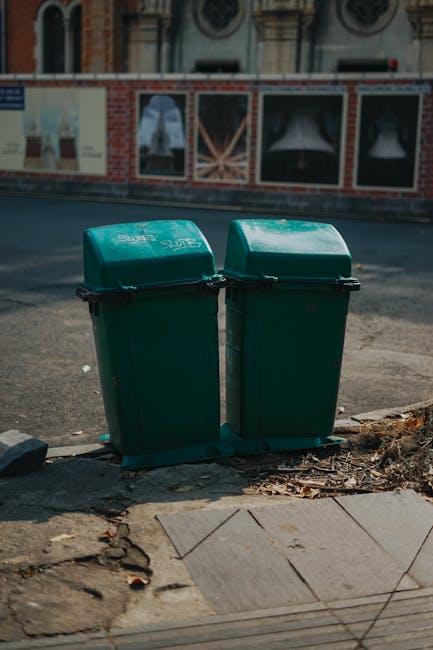 Two green plastic rubbish bins with hinged lids are positioned side by side on a paved sidewalk at the edge of a street, with a small pile of dry leaves and debris around their base. The bins are made of durable moulded plastic with textured matte finishes, featuring black handles on the front for easy opening. Behind the bins, a street surface with asphalt is visible, and across the road, a construction hoarding painted with posters and images partially blocks the view of a brick building with decorative window frames. The lighting suggests daylight, with soft shadows cast by the bins and some glare on their surfaces, indicating a clear or partly cloudy sky. The setting reflects a typical urban environment where private waste collection services, like those offered by rubbishcollectionwestkensington.co.uk, are used to manage refuse outside residential or commercial premises, especially in areas where on-site waste disposal is manually handled before collection.