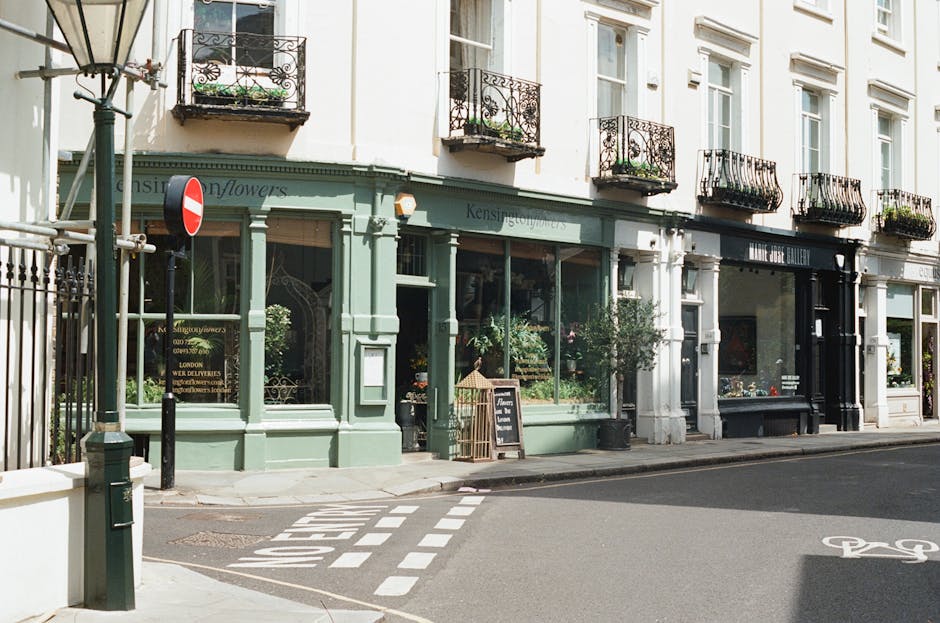A street view showing a row of three- to four-story commercial buildings with residential flats above, featuring balconies with wrought iron railings. The closest building is painted in a pastel green hue with large glass display windows and a black-framed entrance door, indicating a flower shop. Potted plants and flowers are arranged on the sidewalk outside, along with a small A-frame chalkboard sign. To the right, another storefront has a black facade with large display windows, possibly an art gallery or boutique. The sidewalk runs alongside a narrow grey asphalt street with clear road markings for a pedestrian crossing. A traffic sign on a pole is visible on the left side near the corner of the green building. The scene is illuminated by natural daylight, with shadows cast on the pavement. This street scene exemplifies the character of a mixed-use urban area, where shops and residences coexist, and the environment is maintained for pedestrians and local business activity, relating to the need for regular rubbish collection services such as those provided by West Kensington rubbish collection specialists.