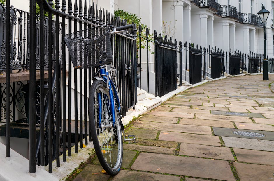 In the foreground, a blue bicycle with a black front basket and silver spokes is locked to a black metal fence that lines a residential street. The fence features vertical bars with pointed tips and decorative elements at the top, running parallel to the paved sidewalk. Behind the fence, there are white-painted stone steps leading up to a row of elegant white terraced houses with black wrought-iron balconies, ornate railings, and traditional lantern-style street lamps mounted on building facades. The sidewalk is constructed from large, uneven stone slabs with narrow gaps filled with moss or dirt, indicating a well-established urban area. The scene is evenly lit with natural daylight, creating a neutral and calm atmosphere typical of a quiet street in West Kensington. This setting subtly relates to private waste handling or on-site clearance by illustrating the typical environment where such services may be applied, with the bike and street environment providing context for everyday urban waste and clutter management, aligning with services like those offered by [COMPANY_NAME] for rubbish collection and clearance in the Kensington and Chelsea area.